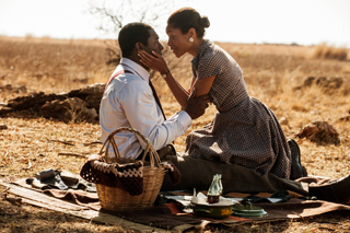 A man and woman sit on a picnic blanket in a dry field, facing each other closely.