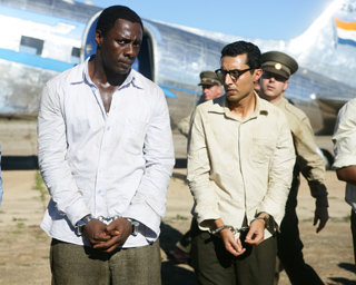 Two men in light-colored shirts stand handcuffed in front of a large airplane.
