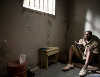 A man in tan clothing sits on a thin mattress in a dim cell, reading a book by light from a barred window.