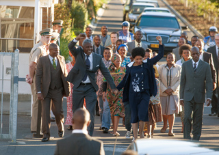 A group of people in formal attire walking along a street with cars parked in the background.