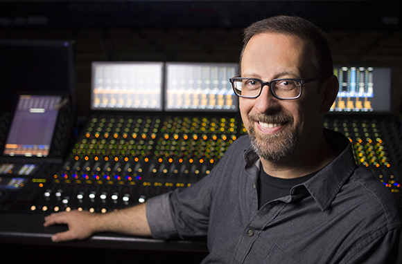 A man with glasses and a goatee smiling in a recording studio in front of a mixing board.