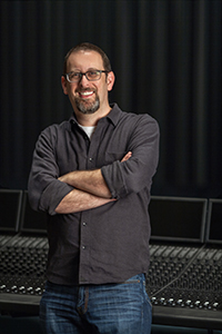 A man with glasses and a goatee stands with arms crossed in front of a large audio mixing console.