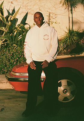 A man in a white hoodie and black pants leans against the front of a red car outdoors.