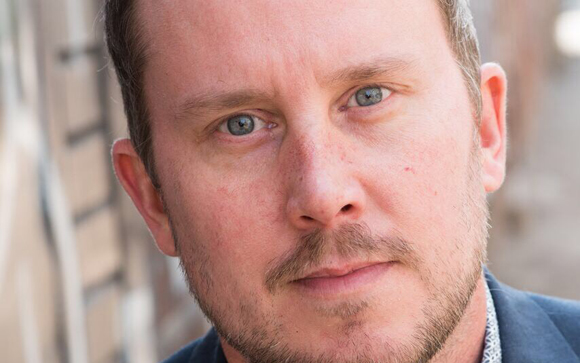 Close-up portrait of a man with light blue eyes and a short beard looking directly at the camera.