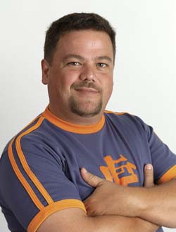 A man with short dark hair and a goatee poses with arms crossed, wearing a purple shirt with orange trim.