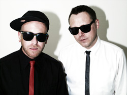 Two men wearing sunglasses, one in a black shirt with a red tie, the other in a white shirt with a black tie.