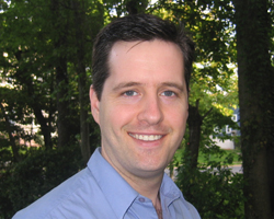 A smiling man with dark hair wearing a light blue button-down shirt outdoors with green trees in the background.