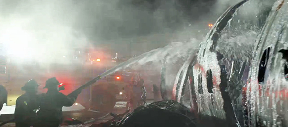 Firefighters aim a high-pressure hose at the charred, metallic wreckage of an airplane at night.