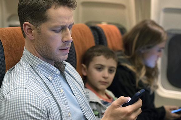 A man in a checkered shirt looks at his smartphone while seated on a plane next to a young boy.
