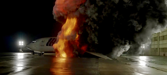 A massive orange and black explosion erupts next to a white airplane on a dark tarmac.