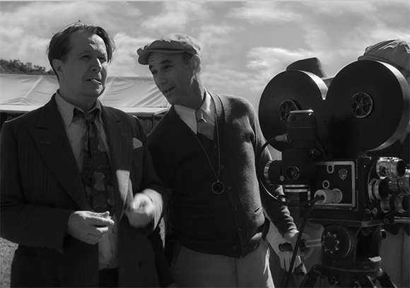 Two men in mid-century attire stand beside a large vintage film camera on a tripod.