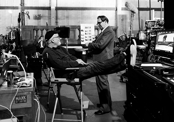 A man in a cap sits in a director's chair while another man in a suit stands talking to him on a film set.