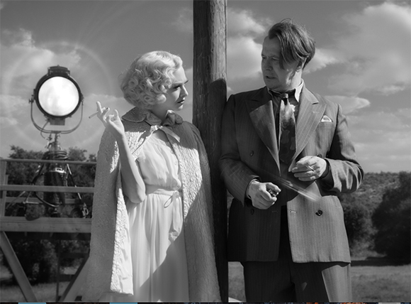 A blonde woman in a light dress and a man in a suit stand next to a wooden pole under a cloudy sky.