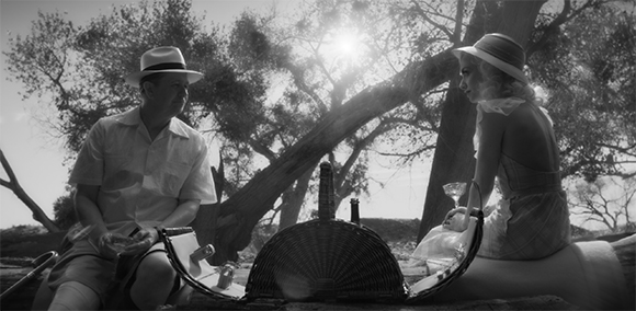 Black and white photo of a man and woman sitting by an open picnic basket under trees in sunlight.