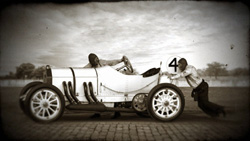 Two men in vintage attire with a white antique racing car numbered 4 on a dirt track.