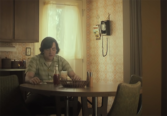 A person with dark hair sits at a wooden dining table in a vintage-style kitchen.