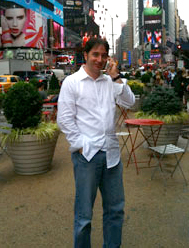 A man in a white button-down shirt and blue jeans stands in a crowded city plaza with bright billboards.