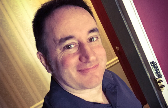A man with short dark hair smiling at the camera in an indoor setting.