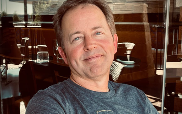 A man with short brown hair smiling at the camera in a brightly lit room.