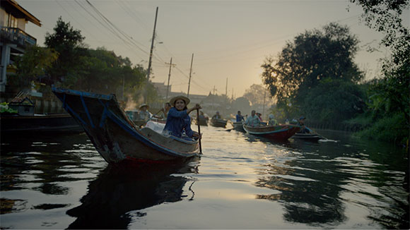 A person in a blue shirt and straw hat rows a small wooden boat through a canal lined with trees and buildings.