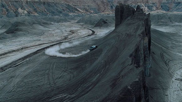 A dark car speeds across a dusty, winding path in a vast, grey mountainous desert terrain.