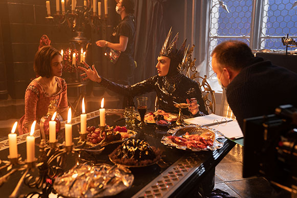 Actors in period costumes sit at a candlelit table while a crew member films the scene.