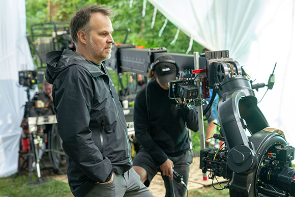 A man in a black jacket stands amidst film cameras and equipment on a production set.