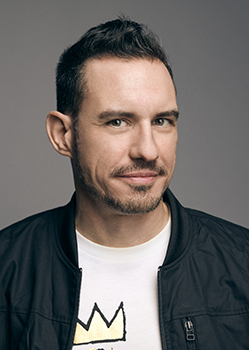 A man with short dark hair and a light beard looks forward against a gray background.