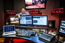 A man sits behind a desk filled with multiple computer monitors, speakers, and audio mixing equipment.