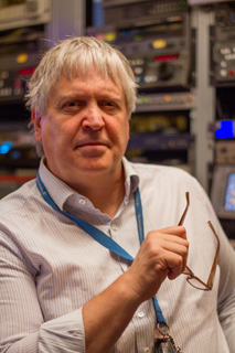A man with grey hair wearing a light striped shirt and a blue lanyard, holding glasses in a server room.