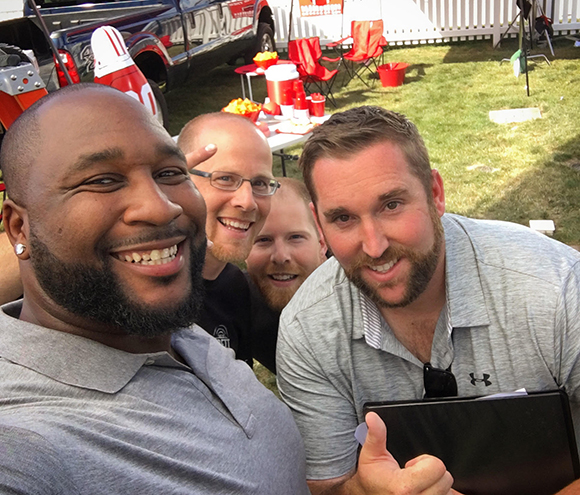 Three men smiling closely together during an outdoor gathering.