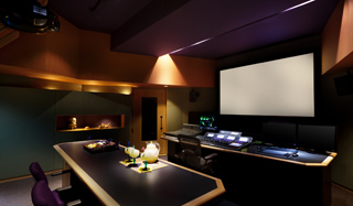 A dark media control room with a large projection screen, dual monitors, and a long desk with equipment.