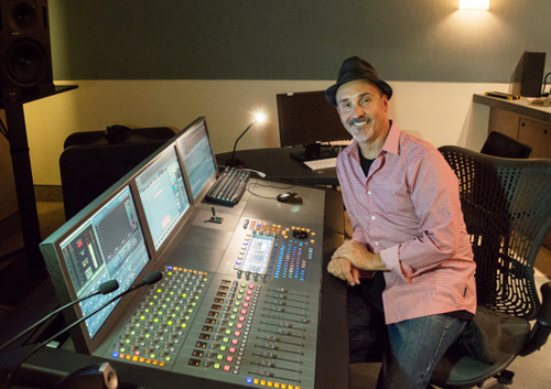 A man wearing a black hat and pink patterned shirt sits at a large professional audio mixing console.