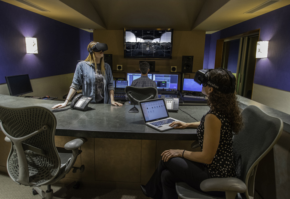 Two people wearing VR headsets sit at a desk with monitors and laptops in a dimly lit room.