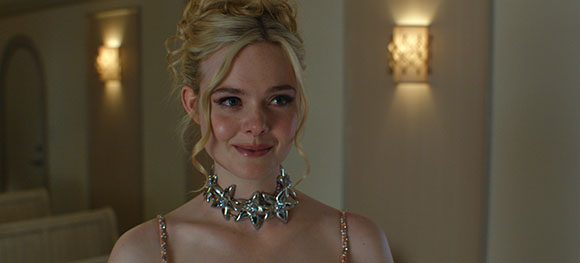 A blonde woman with an updo and a silver statement necklace smiles in a dimly lit room.
