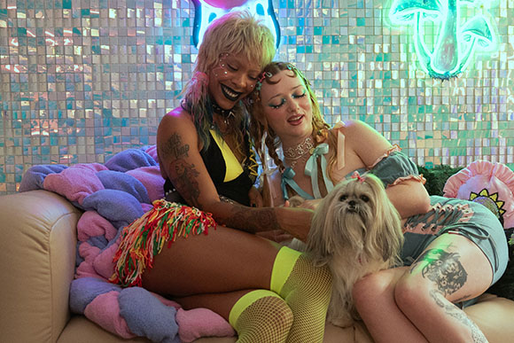 Two women with colorful hair and tattoos sit on a couch with a small white dog against a mosaic background.