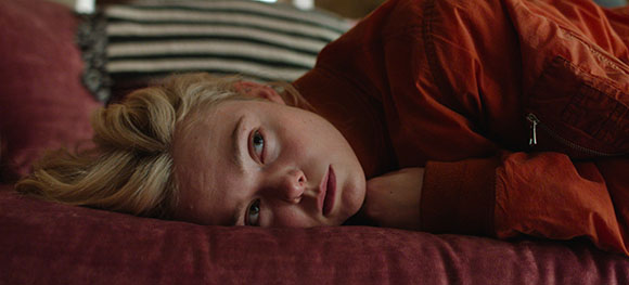 A blonde person in an orange jacket lies on a red cushion with a somber expression.