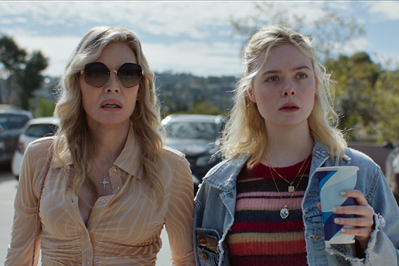 A blonde woman in sunglasses and a blonde woman in a denim jacket look ahead with surprised expressions.