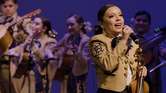 A female mariachi singer sings into a microphone while holding a violin, with band members behind her.