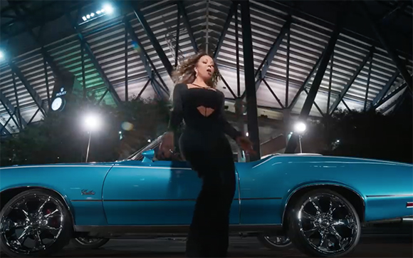 A woman in a black dress leans against a blue vintage convertible under a large metal structure at night.