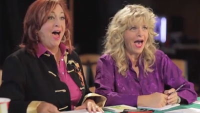 Two women with expressive, joyful facial expressions sitting at a table.