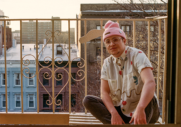A person in a pink beanie and glasses sits on a balcony overlooking city buildings.