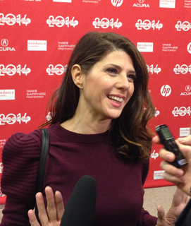 A woman with dark hair smiles while being interviewed in front of a red backdrop with "2014" and "Sundance" logos.
