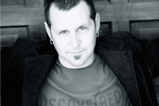 Black and white close-up portrait of a man with short hair and a small goatee leaning against a wooden wall.