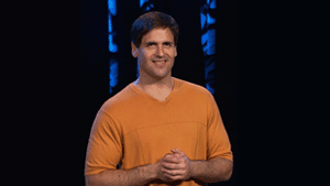 A man with short dark hair wearing an orange v-neck t-shirt, smiling with hands clasped.