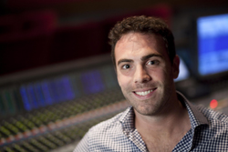 A smiling man in a checkered shirt sitting in front of a large audio mixing console.