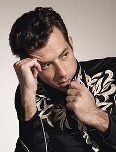 A man with dark hair wearing a black shirt with gold embroidery, resting his chin on his hand.