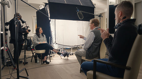 A film crew with cameras and lighting equipment interviews a woman seated in a chair.