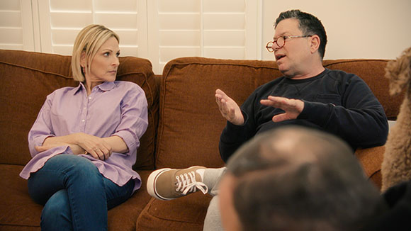 A blonde woman in a purple shirt looks at a man in a dark sweater gesturing while they sit on a brown couch.