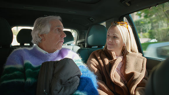 A man in a colorful sweater and a woman in a brown coat sit in the back seat of a vehicle.